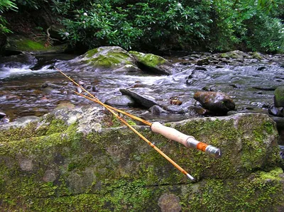 A beautifully crafted fly rod resting on a moss-covered rock with a small, clear stream flowing over rocks in the background.